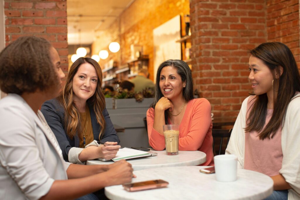 Four women seated at a small round table in a cozy cafe, engaged in a lively conversation. The background shows a warm brick interior with soft lighting, creating an inviting atmosphere.